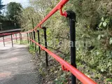 red and black powder coated DDA disability handrail at railway station. Green bushes and trees to the right